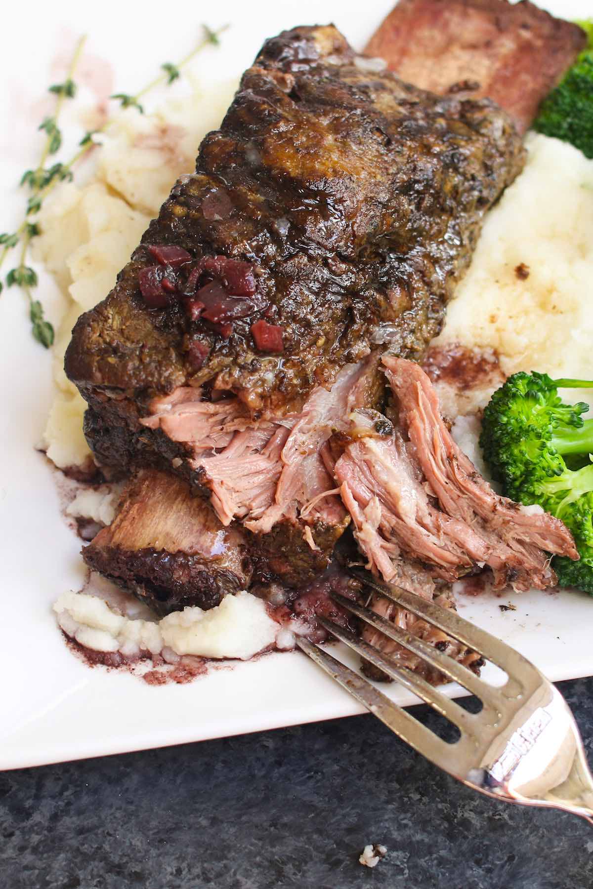 Primer plano que muestra la carne tierna que se cae del hueso de las costillitas de res al vacío con salsa de vino tinto, servidas sobre puré de papas con brócoli al vapor.